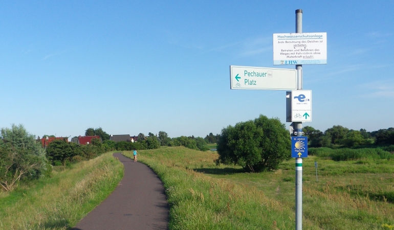 Blick auf den Elberadweg in Pechau entlang des asphaltierten Weges auf des Deichkrone. Vorn ein Hinweisschild auf den Elberadweg und den Pechauer Platz.