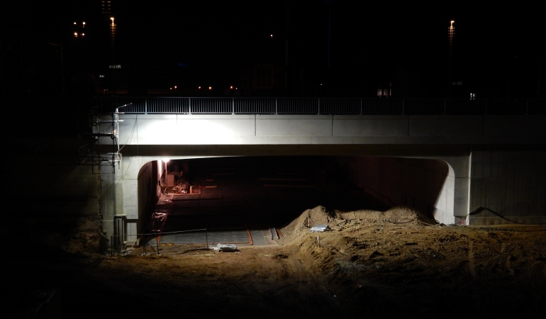 Neubau der westlichen Eisenbahnbrücke mit darunterliegender Tunneldecke im Dunkeln, von Bauscheinwerfern erleuchtet.