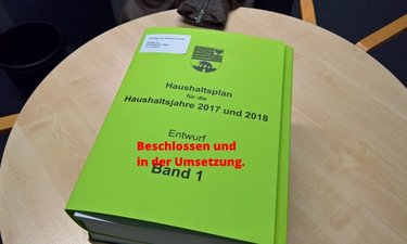 Der erste Band des Haushaltsentwurf auf einem Tisch mit der einmontierten Schrift - Beschlossen und wird umgesetzt.