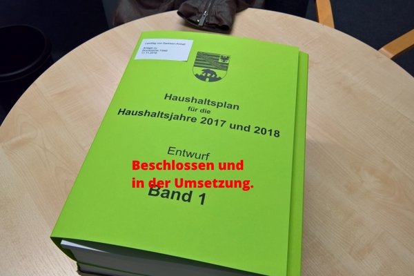 Der erste Band des Haushaltsentwurf auf einem Tisch mit der einmontierten Schrift - Beschlossen und wird umgesetzt.