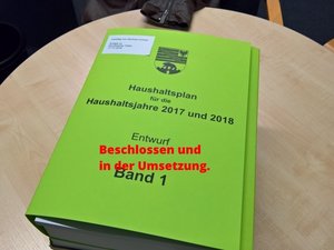 Der erste Band des Haushaltsentwurf auf einem Tisch mit der einmontierten Schrift - Beschlossen und wird umgesetzt.