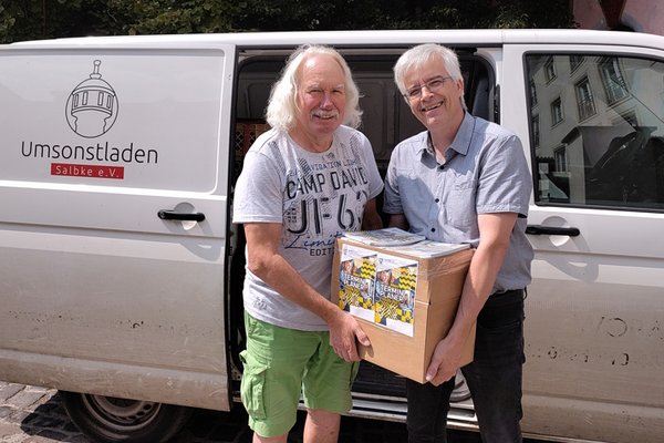 Olaf Meister (rechts) übergibt 2 Klassensätze Terminplaner des Landtages in einem Karton an Ingo Block vom Umsonstladen Salbke e. V. Im Hintergrund der Transporter des Vereins.