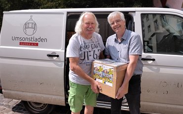 Olaf Meister (rechts) übergibt 2 Klassensätze Terminplaner des Landtages in einem Karton an Ingo Block vom Umsonstladen Salbke e. V. Im Hintergrund der Transporter des Vereins.