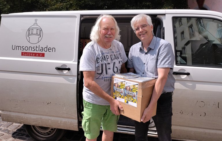 Olaf Meister (rechts) übergibt 2 Klassensätze Terminplaner des Landtages in einem Karton an Ingo Block vom Umsonstladen Salbke e. V. Im Hintergrund der Transporter des Vereins.