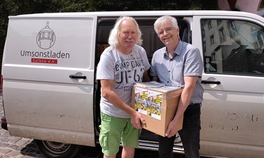 Olaf Meister (rechts) übergibt 2 Klassensätze Terminplaner des Landtages in einem Karton an Ingo Block vom Umsonstladen Salbke e. V. Im Hintergrund der Transporter des Vereins.