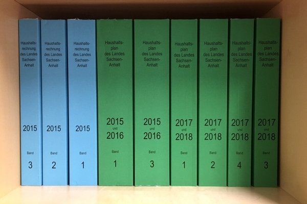 Die Bände der Landeshaushalte von 2015 (blau), 2015/16 und 2017/18 (beide grün) stehend in einem offenen Holzschrank.